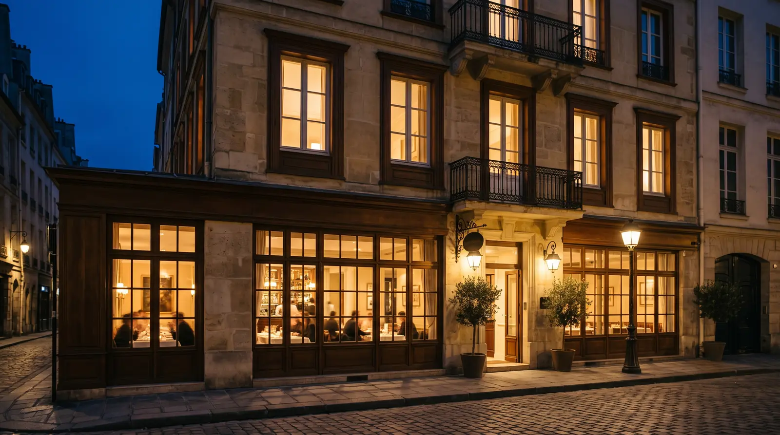 Façade d'un hôtel-restaurant parisien illuminée à la nuit tombée, salle animée derrière les vitrines et rue pavée.